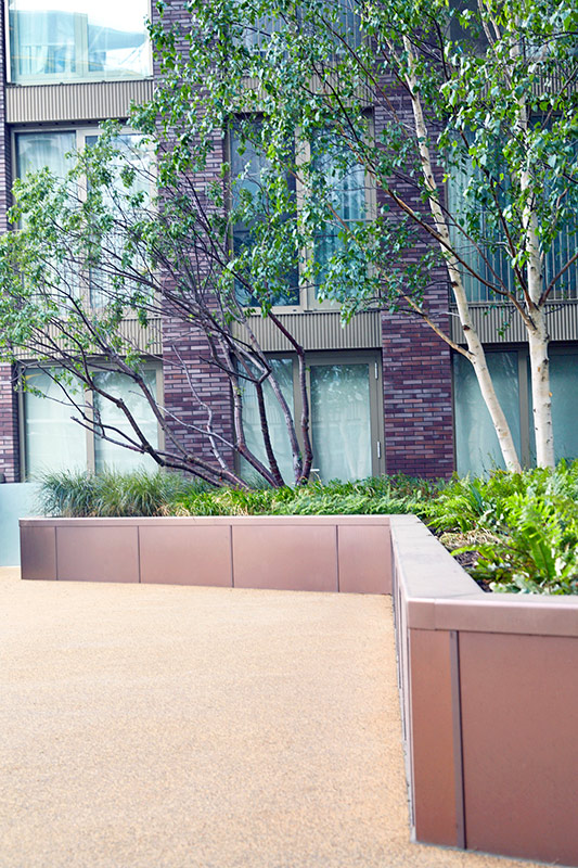 PVD coloured stainless steel in sandblasted Bronze, planters complementing the building materials. In shade the Bronze PVD has a cool tone. Embassy Gardens, London, UK. PVD coloured stainless steel in sandblasted Bronze, planters complementing the building materials. In shade the Bronze PVD has a cool tone. Embassy Gardens, London, UK.