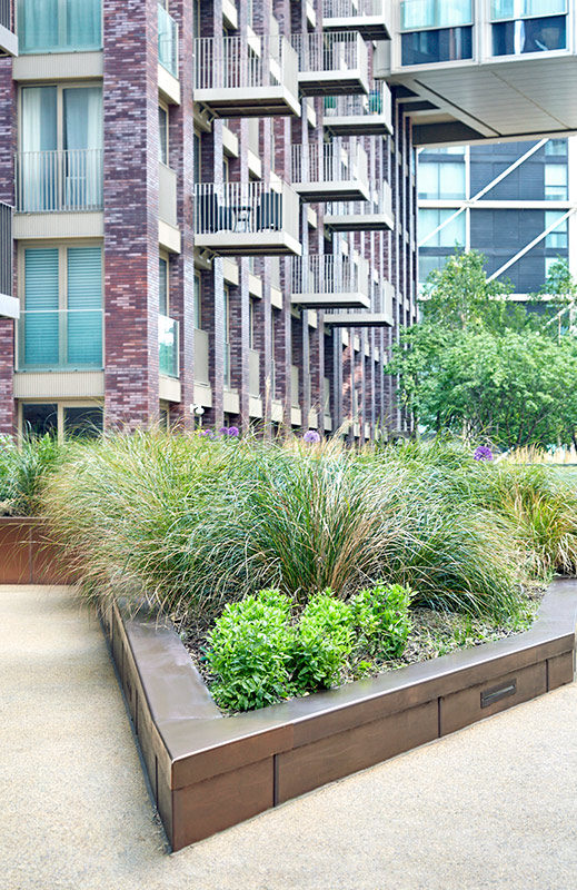 PVD coloured stainless steel in sandblasted Bronze, sharp angled corner section of planter. Embassy Gardens, London, UK. This image was taken in the shade and the Bronze shows as a cool tone. PVD coloured stainless steel in sandblasted Bronze, sharp angled corner section of planter. Embassy Gardens, London, UK. This image was taken in the shade and the Bronze shows as a cool tone.