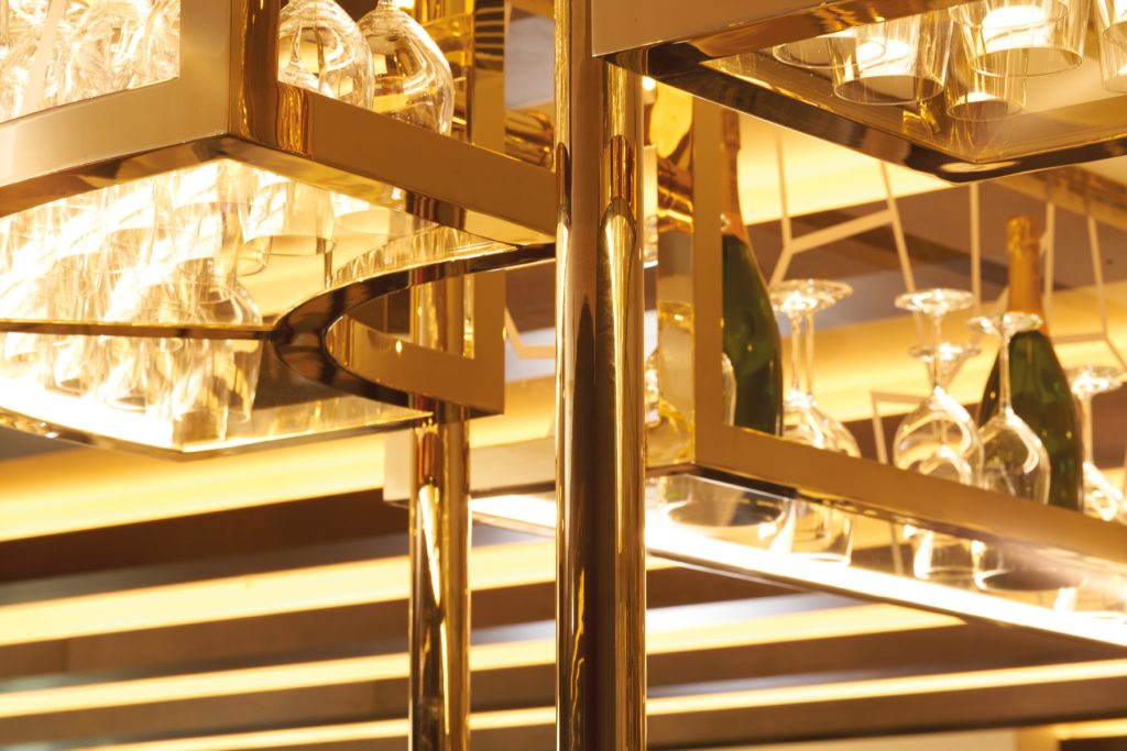 A close-up of the bar metalwork in Champagne Mirror PVD which has the look of a brass finish. The glass rack design combines rectilinear edges with curved profiles and large circular tube for uprights. The Devonshire Club Hotel, Interior Design: March & White, PVD coloured stainless steel: Double Stone Steel Europe Ltd