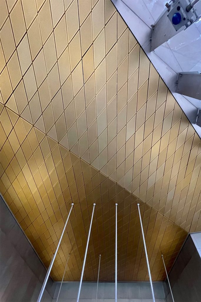 Showing the gold, or brass-coloured, anodised aluminium diagonally-set ceiling panels to the Wood Lane Underground Station, London. Architectural design by Ian Ritchie Architects. Ceiling by Double Stone Steel Europe Ltd.
